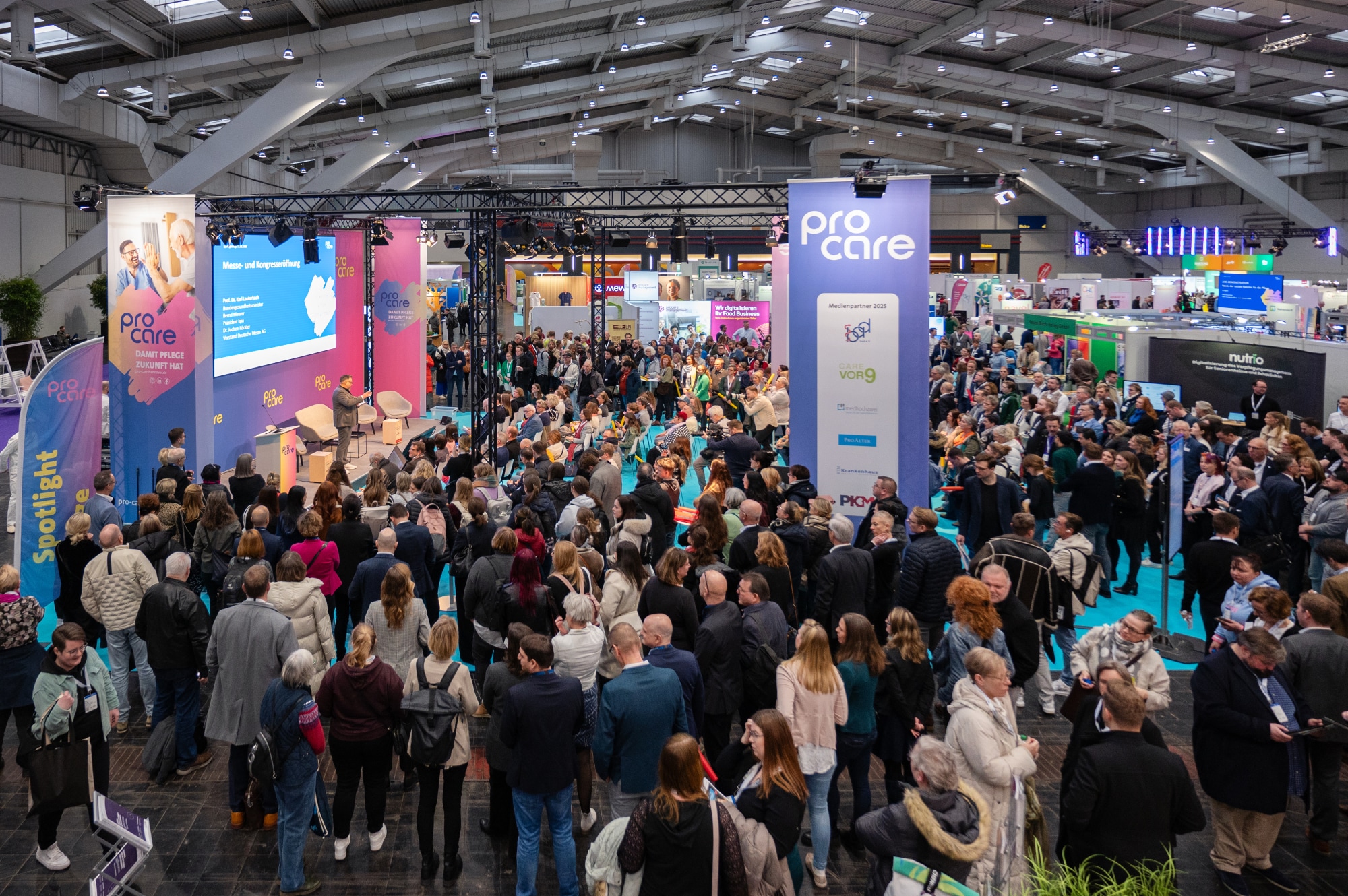 Eine große Gruppe von Menschen steht in einer Messehalle an der Vortragsbühne der Pro Care 